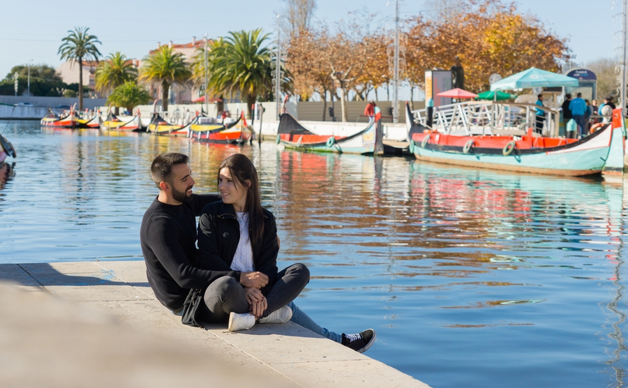 Young couple in love looking at each other while smiling and sitting on the ground near the water canal in Aveiro, and in the background the boats on a sunny day.
