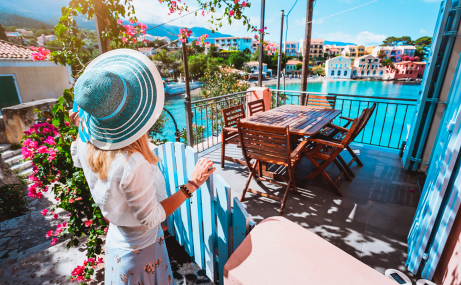 Female tourist with blue sun hat staying in Assos village in front of cozy veranda and admiring turquoise colored bay of Mediterranean sea and beautiful colorful houses of in Kefalonia, Greece