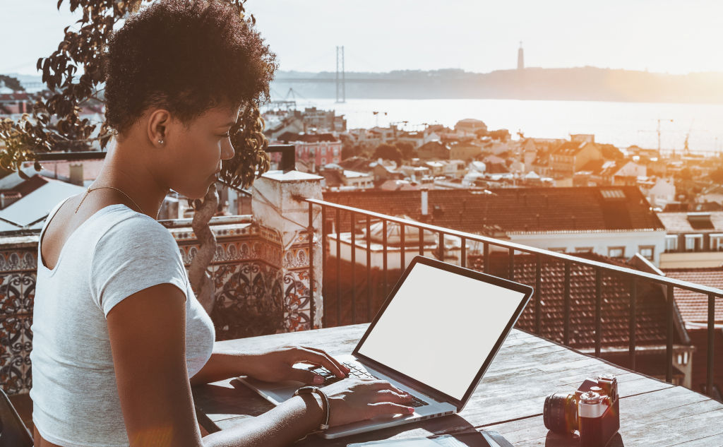 Young appealing biracial female photographer sitting on balcony of Lisbon hotel with stunning bright cityscape in background and working with laptop