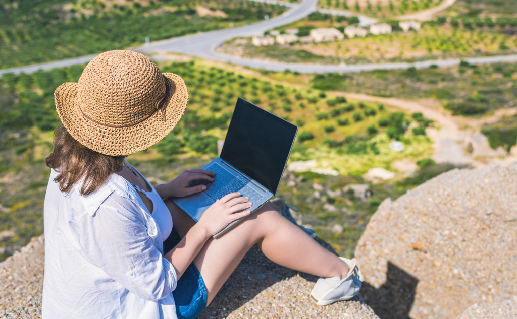 Woman freelancer in tourist clothes and straw hat with a laptop on top of a mountain.
